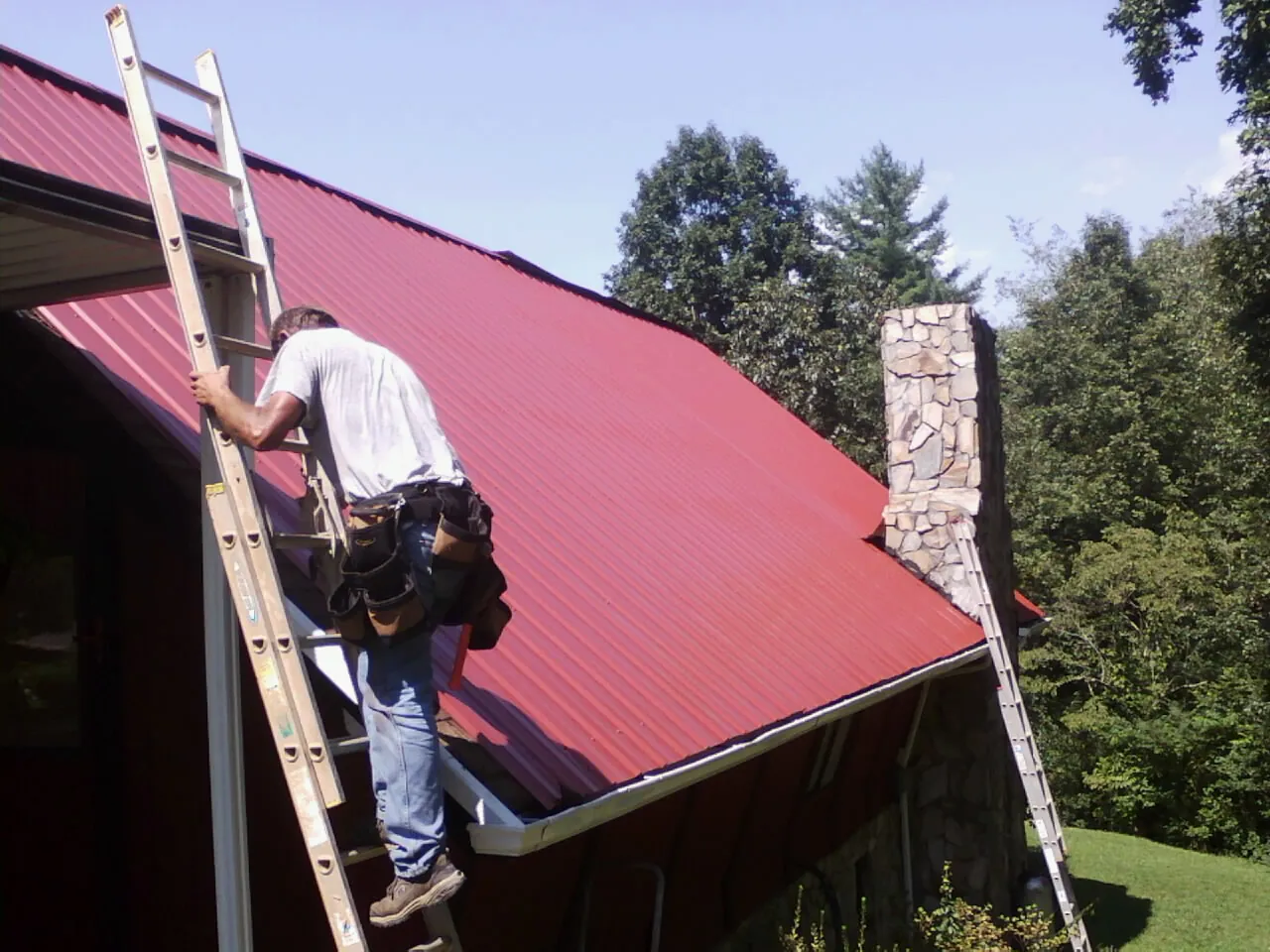 Roofer on a ladder beside a red metal roof and stone chimney — Soffit & Fascia Repair by Roofing Brothers in Zionville